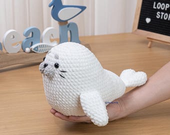 May include: A handmade white crochet seal plush toy with gray accents on the nose and whiskers. The seal is held in a person's hand. The background includes beach-themed decor and a sign that says "THE LOOP STOP".