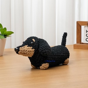 May include: A crocheted dachshund dog figurine in black and tan. The handmade dog has a black body, tan snout, paws, and ears, and a small, upright tail. The dog is sitting on a wooden surface.