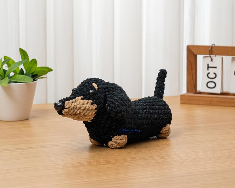 May include: A crocheted dachshund dog figurine in black and tan. The handmade dog has a black body, tan snout, paws, and ears, and a small, upright tail. The dog is sitting on a wooden surface.