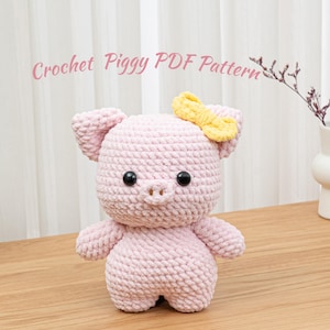 Puede incluir: Un cerdo rosa hecho a mano con un lazo amarillo. El cerdo tiene ojos de botón negros y un pequeño hocico. El texto "Crochet Piggy PDF Pattern" está escrito en rosa en cursiva encima del cerdo.
