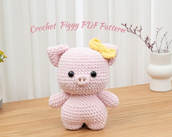 May include: A handmade pink crochet pig with a yellow bow. The pig has black button eyes and a small snout. The text "Crochet Piggy PDF Pattern" is written in pink cursive above the pig.