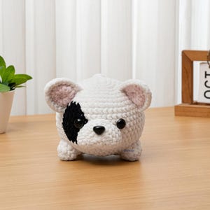 May include: A handmade, crocheted dog figurine in white with a black patch around one eye and black nose. The dog has small, rounded ears with pink inner details. The toy is sitting on a wooden surface.