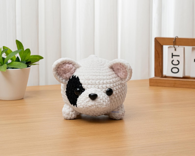 May include: A handmade, crocheted dog figurine in white with a black patch around one eye and black nose. The dog has small, rounded ears with pink inner details. The toy is sitting on a wooden surface.