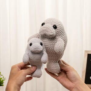 May include: Two light gray crocheted manatee plush toys, one larger than the other, featuring rounded bodies, small side fins, flat tail flukes, and black plastic safety eyes. The textured yarn creates a soft, uniform appearance for both.