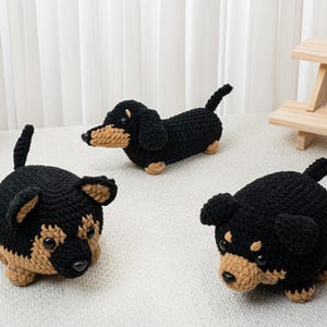 May include: Three crocheted dachshund dog figurines in black and tan. One is lying down, and two are sitting up. The dogs have black bodies, tan faces, ears, and paws. The background is a white surface and a white curtain.