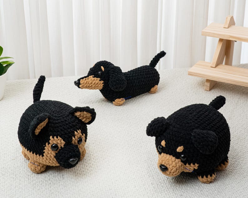 May include: Three crocheted dachshund dog figurines in black and tan. One is lying down, and two are sitting up. The dogs have black bodies, tan faces, ears, and paws. The background is a white surface and a white curtain.
