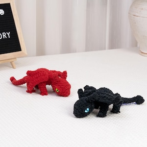 May include: Two crocheted dragon toys. One is red with yellow eyes, the other is black with blue eyes. The toys are lying on a white surface.