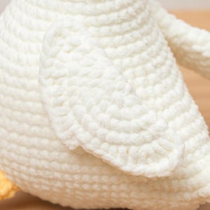 May include: A white crocheted stuffed animal featuring a rounded body and a prominent wing section. The texture is created with dense, uniform yarn loops. A small portion of a yellow crocheted foot is visible at the bottom left of the object.