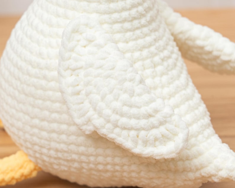 May include: A white crocheted stuffed animal featuring a rounded body and a prominent wing section. The texture is created with dense, uniform yarn loops. A small portion of a yellow crocheted foot is visible at the bottom left of the object.