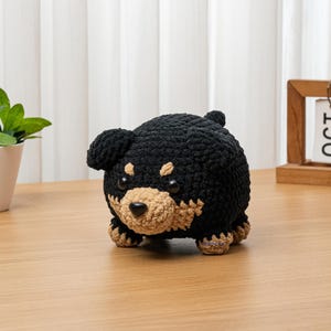 May include: A handmade crochet dog figurine in black and tan. The plush toy has a rounded body, small ears, and a tan snout. The dog is sitting on a wooden surface. The background includes a small plant in a white pot and a wooden calendar.