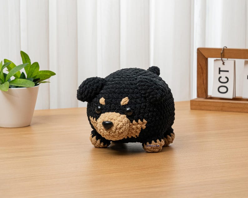 May include: A handmade crochet dog figurine in black and tan. The plush toy has a rounded body, small ears, and a tan snout. The dog is sitting on a wooden surface. The background includes a small plant in a white pot and a wooden calendar.