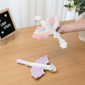 May include: Two handmade crochet dragon toys. One dragon is white with pastel rainbow-colored wings, and the other is white with solid light purple wings. Both feature small black eyes, textured bodies, and detailed wing structures made of yarn.