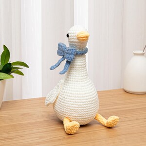May include: A white crocheted goose toy with a rounded body, a long neck, and a yellow beak and feet. The goose features black bead eyes and wears a light blue crocheted bow tied around its neck. The texture of the yarn is visible throughout.