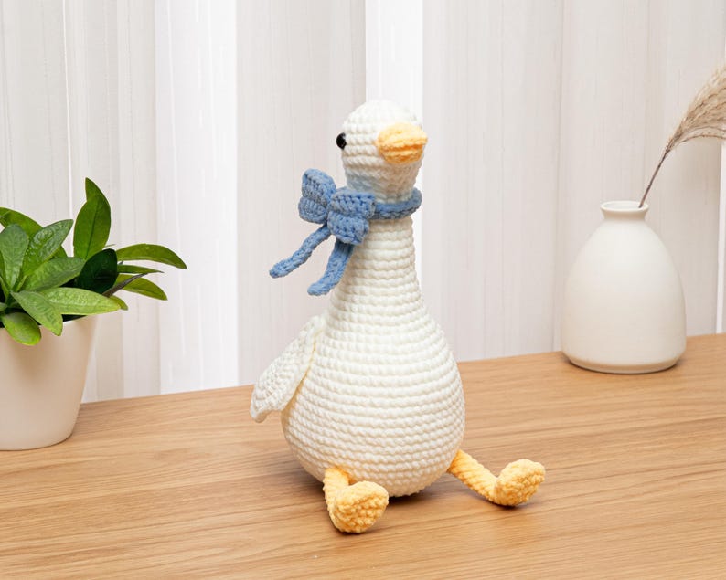 May include: A white crocheted goose toy with a rounded body, a long neck, and a yellow beak and feet. The goose features black bead eyes and wears a light blue crocheted bow tied around its neck. The texture of the yarn is visible throughout.