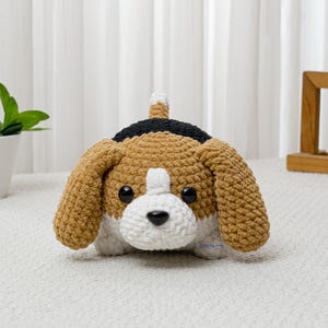 May include: A handmade crochet beagle dog with brown, black, and white details. The plush toy has large floppy ears, a white muzzle, and a black nose. The dog is sitting on a white surface.
