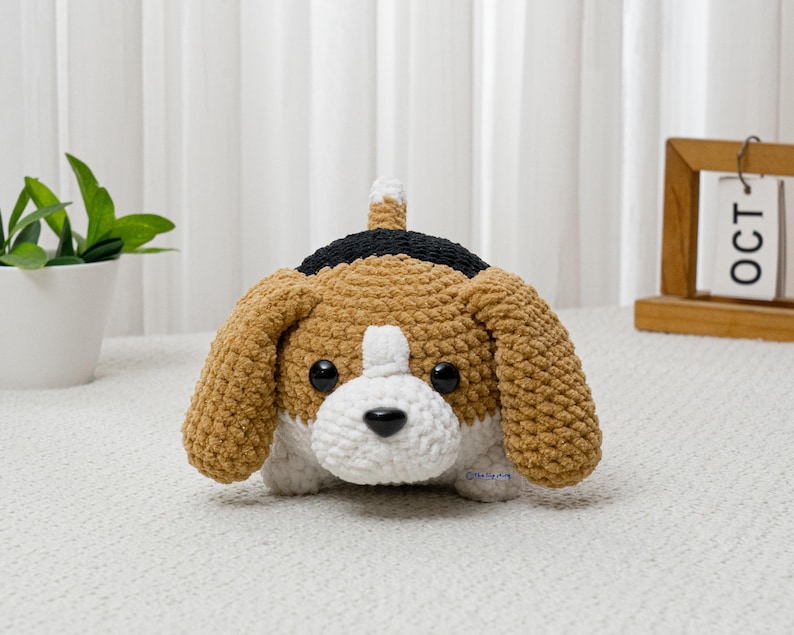 May include: A handmade crochet beagle dog with brown, black, and white details. The plush toy has large floppy ears, a white muzzle, and a black nose. The dog is sitting on a white surface.