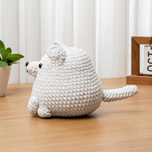 May include: A handmade, white crocheted polar bear toy with black bead eyes and a small tail. The bear is sitting on a wooden surface. A small green plant in a white pot and a wooden calendar are in the background.