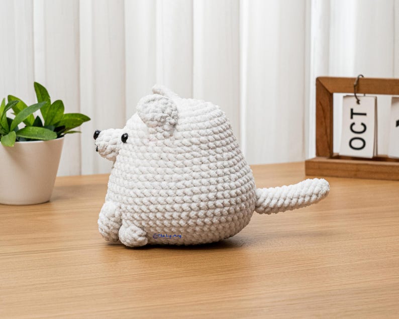 May include: A handmade, white crocheted polar bear toy with black bead eyes and a small tail. The bear is sitting on a wooden surface. A small green plant in a white pot and a wooden calendar are in the background.