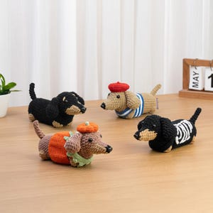 May include: Four crocheted dachshund figurines. One is solid black with tan accents, one is tan with a blue and white striped shirt and red beret, one is brown wearing a pumpkin hat and orange sweater, and one is black with a white skeleton ribcage design.