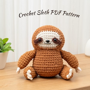 Könnte beinhalten: Eine gehäkelte Faultierpuppe in Braun-, Creme- und Dunkelbrauntönen, mit einem lächelnden Gesicht und kleinen schwarzen Augen. Der Text "Crochet Sloth PDF Pattern" ist über dem Faultier zu sehen. Das Faultier sitzt auf einer Holzoberfläche.