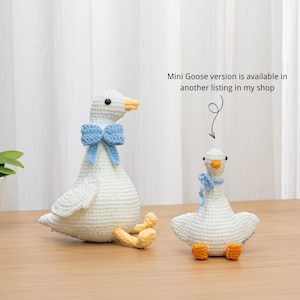 May include: Two white crocheted goose plush toys with orange feet and beaks. The larger goose wears a blue bow, while the smaller goose wears a blue scarf. Text reads: Mini Goose version is available in another listing in my shop.