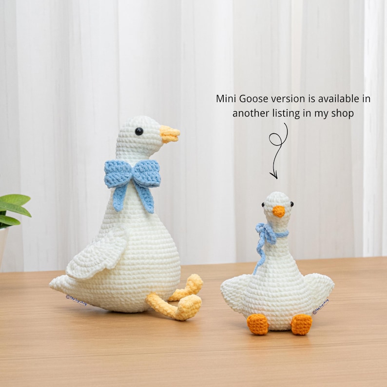 May include: Two white crocheted goose plush toys with orange feet and beaks. The larger goose wears a blue bow, while the smaller goose wears a blue scarf. Text reads: Mini Goose version is available in another listing in my shop.