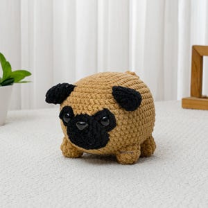 May include: A handmade crochet pug dog with a tan body, black ears, and a black snout. The pug is sitting on a white surface. The dog has large black eyes and small, stubby legs. A cute, cuddly, and soft plush toy.