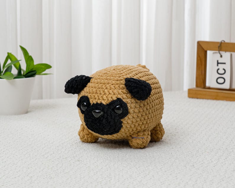 May include: A handmade crochet pug dog with a tan body, black ears, and a black snout. The pug is sitting on a white surface. The dog has large black eyes and small, stubby legs. A cute, cuddly, and soft plush toy.