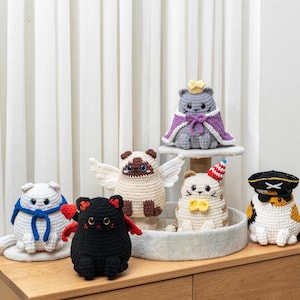 Puede incluir: Una colección de figuras de gatos de ganchillo hechas a mano en varios colores y diseños. Los gatos están adornados con accesorios como una corona, alas y un sombrero de pirata. Las figuras se exhiben sobre una superficie de madera.