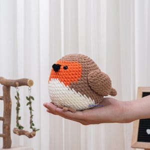 Pode incluir: Um pássaro de crochet feito à mão, com uma forma arredondada. O pássaro tem cabeça e asas castanhas, peito laranja, barriga branca e bico preto. O pássaro é segurado em uma mão.