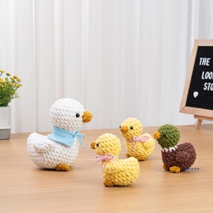 May include: A group of handmade crochet ducks in different sizes and colours. The largest duck is white with a blue scarf, while the smaller ones are yellow and brown. The ducks have orange beaks and feet, and some have pink bows.