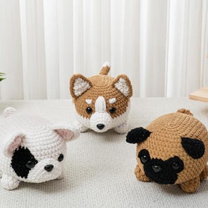 May include: Three crocheted dog figures in white, brown, and tan. The white dog has a black eye patch, the brown dog has a white chest and face, and the tan dog is a pug with black features. The dogs are on a light surface.