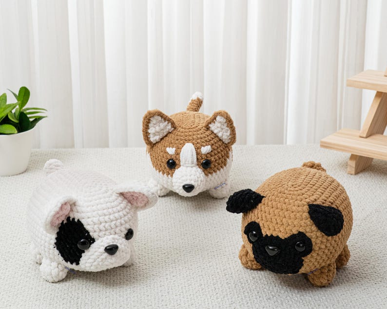 May include: Three crocheted dog figures in white, brown, and tan. The white dog has a black eye patch, the brown dog has a white chest and face, and the tan dog is a pug with black features. The dogs are on a light surface.