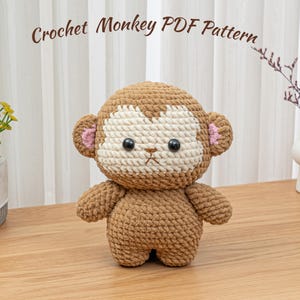 Può includere: Una scimmia all'uncinetto fatta a mano, di colore marrone con testa beige e viso bianco. La scimmia ha occhi neri e orecchie rosa. Il testo "Crochet Monkey PDF Pattern" è visibile nella parte superiore dell'immagine.