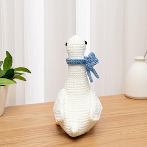 May include: A white crocheted goose toy featuring a textured yarn surface, small black bead eyes, and a light blue crocheted scarf tied around its neck. The goose is crafted with visible stitch patterns and has a rounded body and wings.