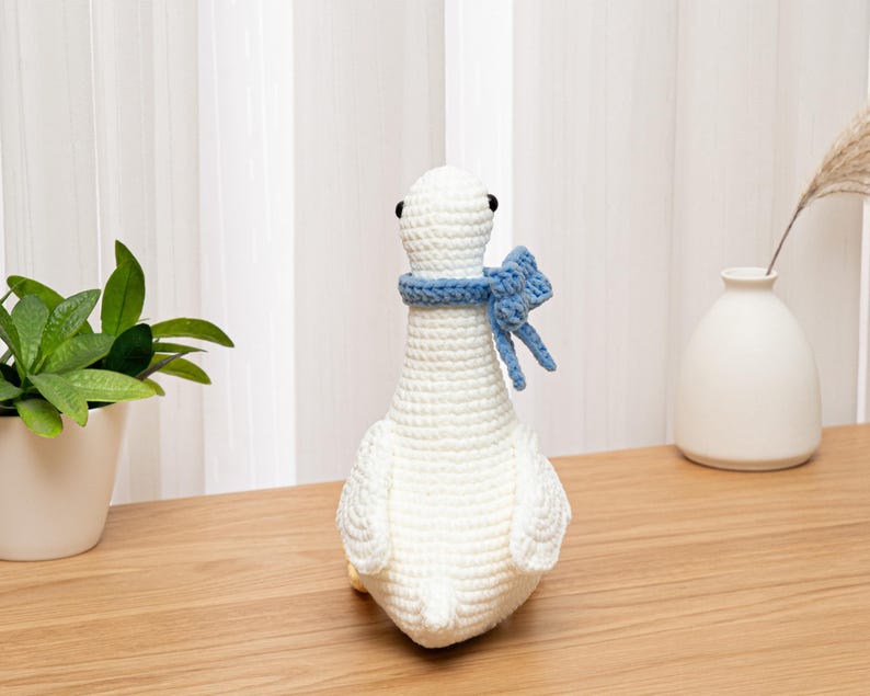 May include: A white crocheted goose toy featuring a textured yarn surface, small black bead eyes, and a light blue crocheted scarf tied around its neck. The goose is crafted with visible stitch patterns and has a rounded body and wings.