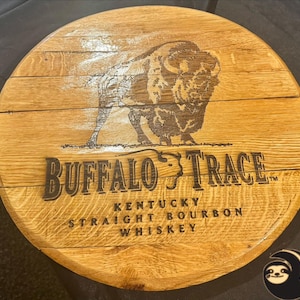 May include: A circular wooden sign featuring the words "Buffalo Trace" and a bison image. The sign also includes the text "Kentucky Straight Bourbon Whiskey". The wood has a natural, light brown colour.
