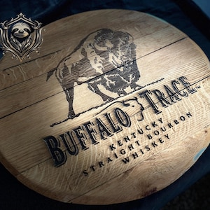 May include: A circular wooden sign featuring the text "Buffalo Trace Kentucky Straight Bourbon Whiskey" in black. A detailed etching of a bison is above the text. The sign is made of light-coloured wood.