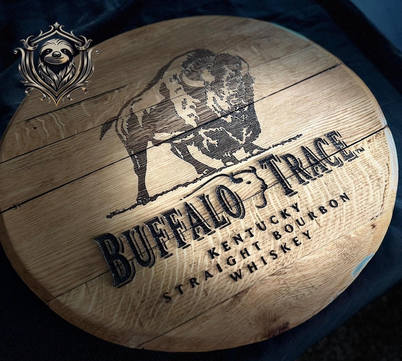 May include: A circular wooden sign featuring the text "Buffalo Trace Kentucky Straight Bourbon Whiskey" in black. A detailed etching of a bison is above the text. The sign is made of light-coloured wood.