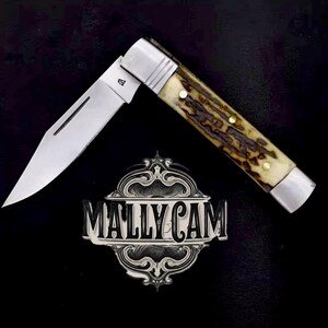 May include: A stainless steel folding knife with a partially open blade. The handle features a brown and cream-colored pattern with brass rivets. The knife is set against a black background, with a decorative emblem that reads "MALLY CAM".