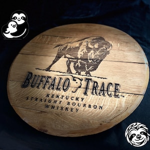May include: A circular wooden sign displaying the Buffalo Trace logo and text. The sign features a detailed image of a bison and the words "Buffalo Trace Kentucky Straight Bourbon Whiskey". The wood has a natural grain and a warm, brown colour.