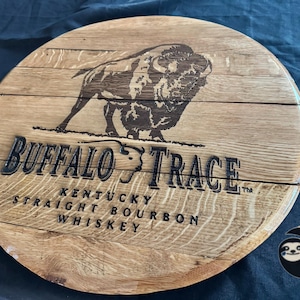 May include: Circular wooden sign featuring the text "Buffalo Trace Kentucky Straight Bourbon Whiskey" and a bison illustration. The sign is made of light brown wood with dark brown text and image.