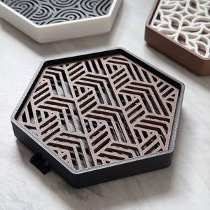 Peut inclure: Ensemble de dessous de verre hexagonaux aux motifs et couleurs variés. L'un présente un motif géométrique marron dans un cadre noir. Un autre affiche un motif tourbillon gris dans un cadre blanc. Un troisième a un motif de feuilles blanches dans un cadre marron.