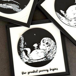 Könnte beinhalten: Schwarz gerahmte Kunstwerke mit einem Graustufen-Ultraschallbild eines Babys in einem schwarzen Kreis. Das Kunstwerk hat einen weißen Rand und den Text "Our greatest journey begins" in einer Schriftart.
