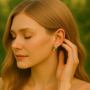 Op de afbeelding: Een close-up van een persoon die een gouden bloemenknopoorbel draagt. De oorbel heeft een delicaat ontwerp, met een klein bloem- of bladmotief. De persoon heeft een lichte huidskleur en lang blond haar.