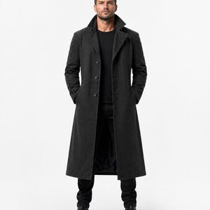 May include: A full-length, dark gray coat with a classic design. The coat features a notched collar, button closure, and front pockets. The model is wearing a black t-shirt, black pants, and black boots. The coat is a stylish outerwear option.