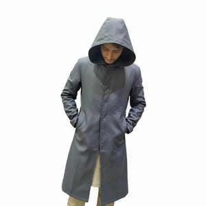 May include: A long, grey hooded coat with a button closure and pockets. The coat is made of a smooth material and has a tailored fit. The hood is large and covers the head. The person is wearing a light-coloured shirt and khaki trousers.