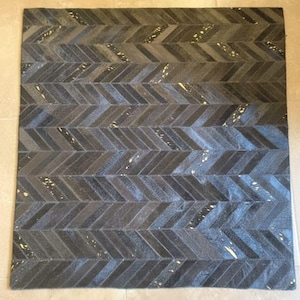 Könnte beinhalten: Ein quadratischer, dunkelgrauer Teppich mit Fischgrätmuster. Der Teppich besteht aus einem strukturierten Material mit Farbvariationen und kleinen, helleren Akzenten. Das Design ist eine sich wiederholende Reihe von Winkellinien.
