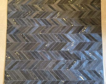 Può includere: Un tappeto quadrato grigio scuro con un motivo a spina di pesce. Il tappeto è realizzato in un materiale strutturato, con variazioni di colore e piccoli accenti più chiari. Il design è una serie ripetuta di linee angolate.
