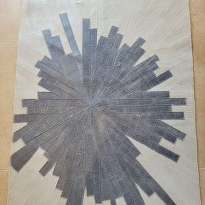 May include: A rectangular rug with a sunburst design. The rug features a central grey burst pattern surrounded by a cream-coloured background. The grey pattern is made of rectangular shapes radiating outwards.